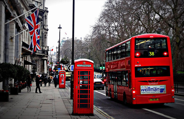 Red double decker bus driving on a London street with UK flags on the building.