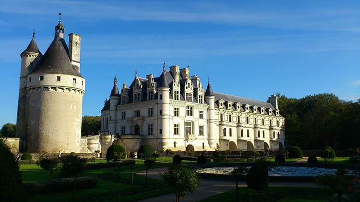 View of Château de Chenonceau, a distinctive French castle with a large central building flanked by round towers, situated beside a manicured garden and a reflective water pond under a clear sky.