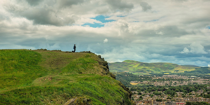 A wide view of Holyrood Park in Edinburgh, Scotland, showing a person standing alone on a grassy cliff edge near the top of a ridge. The foreground is covered in lush green grass and rocky outcrops. Below the cliff, the city of Edinburgh is visible, with clusters of buildings and tree-lined streets. In the background, rolling green hills stretch into the distance beneath a dramatic, partly cloudy sky.