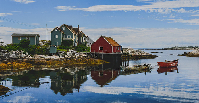 A peaceful coastal scene in a small Canadian fishing village, showing colorful wooden houses—one red and several green—perched on a rocky shoreline. Two small boats float in the calm, reflective water of the harbor. Seaweed-covered rocks line the shore, and the sky above is partly cloudy with patches of blue, adding to the tranquil atmosphere.