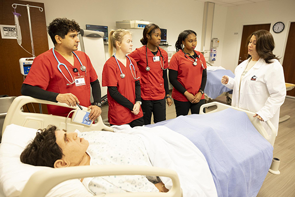 Four NIU nursing students in red scrubs observe and learn from an instructor in a white lab coat during a simulation exercise with a high fidelity medical mannequin in a hospital bed.