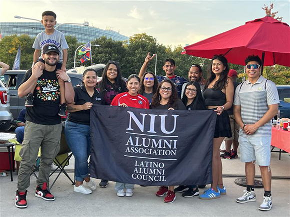 Group of NIU alumni and friends at the Chicago Fire game tailgate, holding an NIU Latino Alumni Council flag