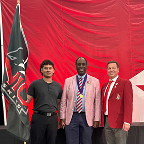 Three men standing in front of a red back drop, one of them is holding a black NIU Huskies flad on a flag pole.